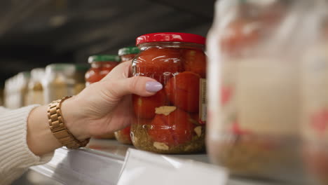close up of shopper hands taking transparent glass jar of tomatoes from supermarket shelf rails in bright grocery aisle highlighting polished nails wrist watch and blurred retail background
