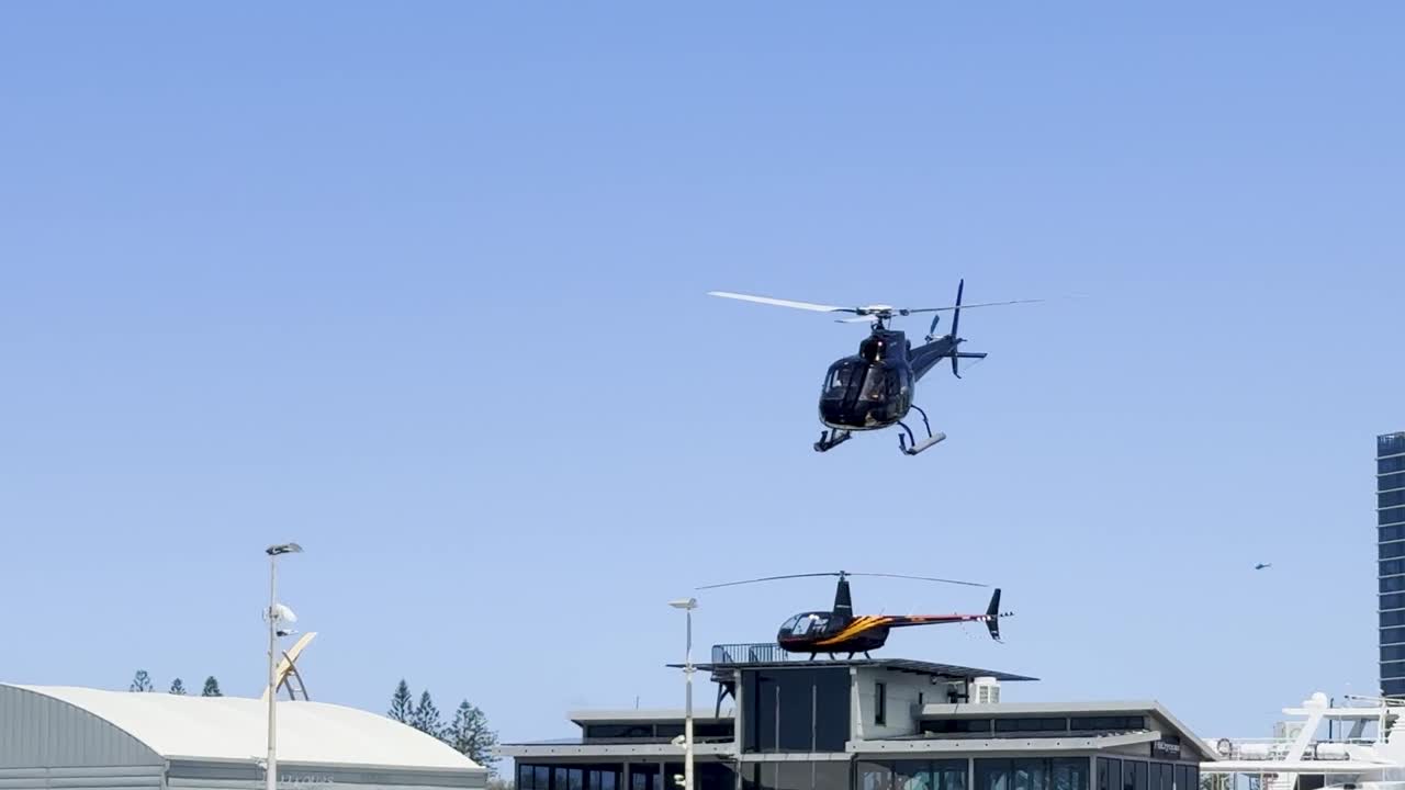 A helicopter hovers above city rooftops with a clear blue sky backdrop.