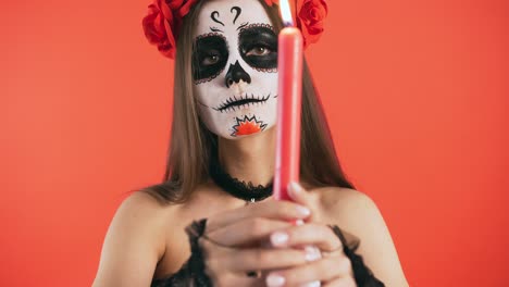 día de los muertos, mujer con maquillaje de cráneo moviendo una vela en círculo y apagándola bruscamente, posando sobre fondo rojo, halloween