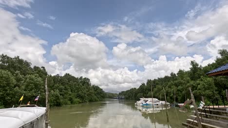 wide-angle daytime pan across a tranquil mangrove river canal with moored speedboats, wooden pier, and lush green forest under partly cloudy skies