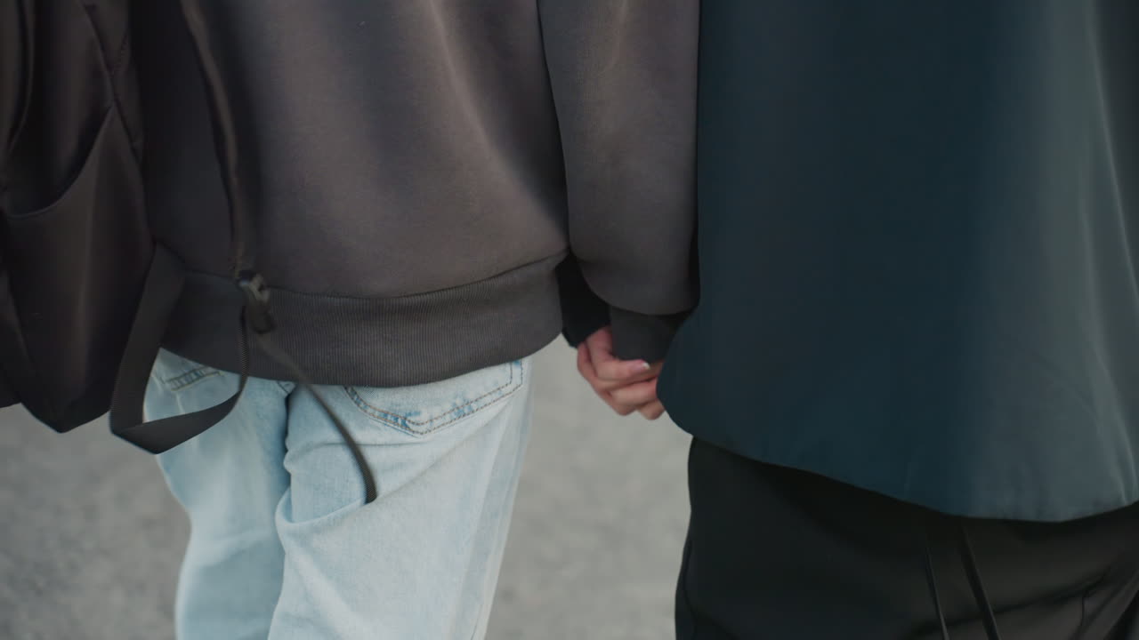 two people holding hands on street, closeup of interlaced fingers with denim and dark jacket, backpack hint and passerby blur suggest student and artist roles, tender connection in soft overcast