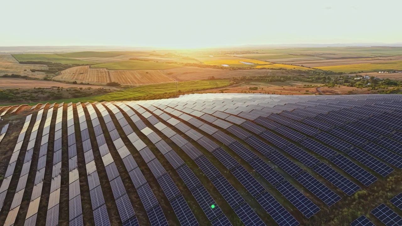 Solpaneler dækker land i et stort felt, mens solen går ned i baggrunden, og viser rækker af paneler, der reflekterer lys