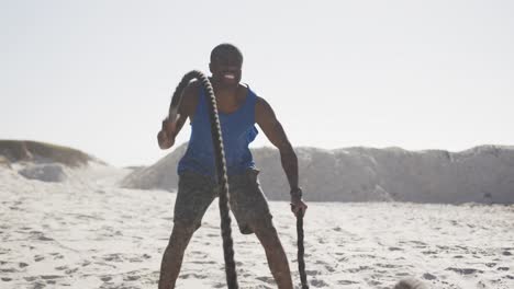 Lächelnder-Afroamerikanischer-Mann,-Der-Draußen-Am-Strand-Mit-Kampfseilen-Trainiert