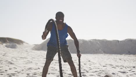 hombre afroamericano sonriente haciendo ejercicio con cuerdas de lucha al aire libre en la playa