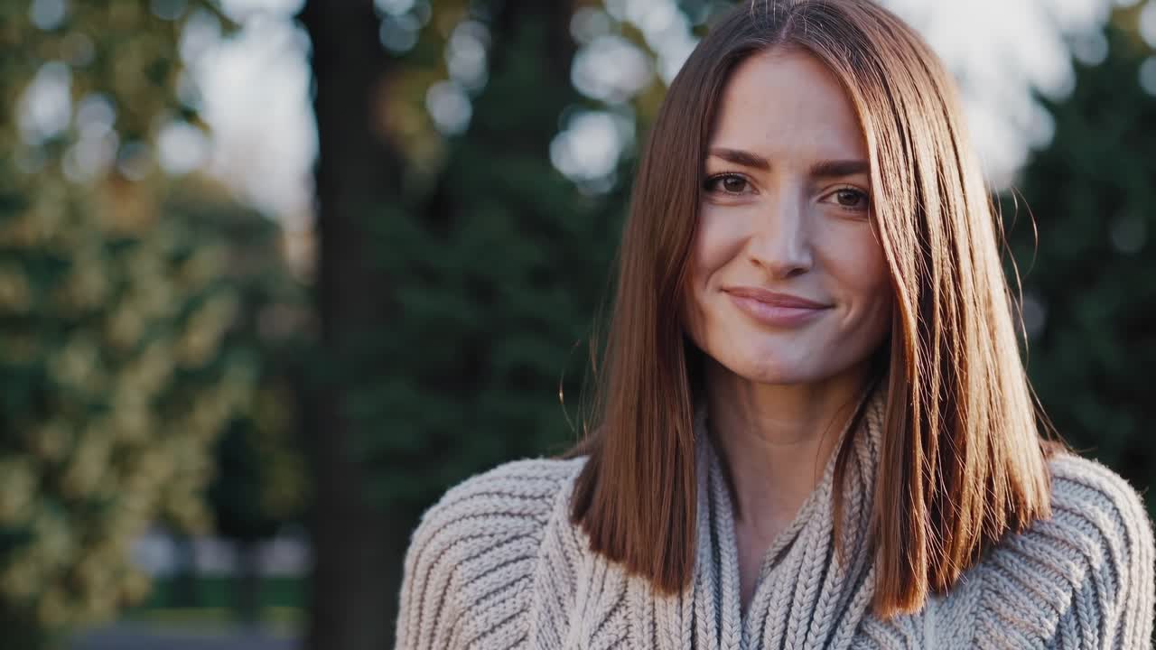 Serene autumn park portrait featuring young woman smiling warmly, enjoying golden sunlight while standing amid falling leaves, radiating natural beauty and peaceful mood