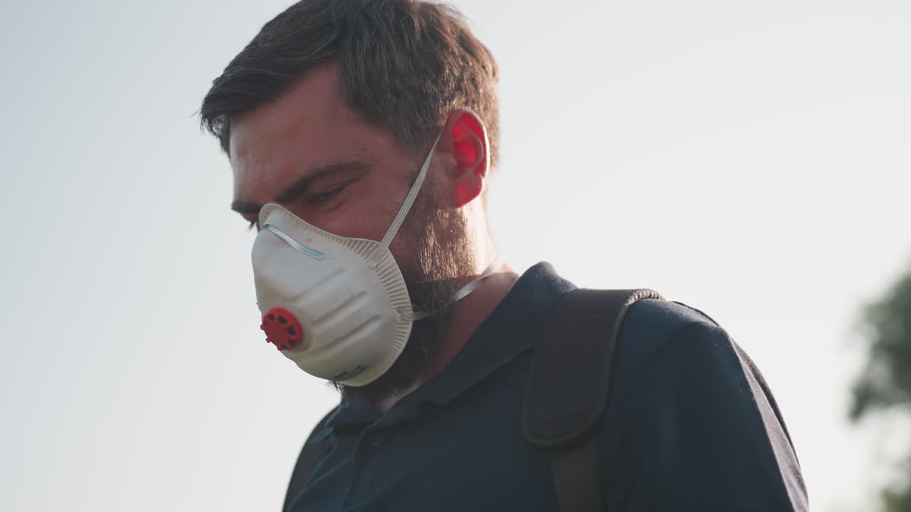 man wearing protective mask performing fumigation in field with sunlight backlight creating flare, serious expression, environmental pest control activity during daytime outdoors