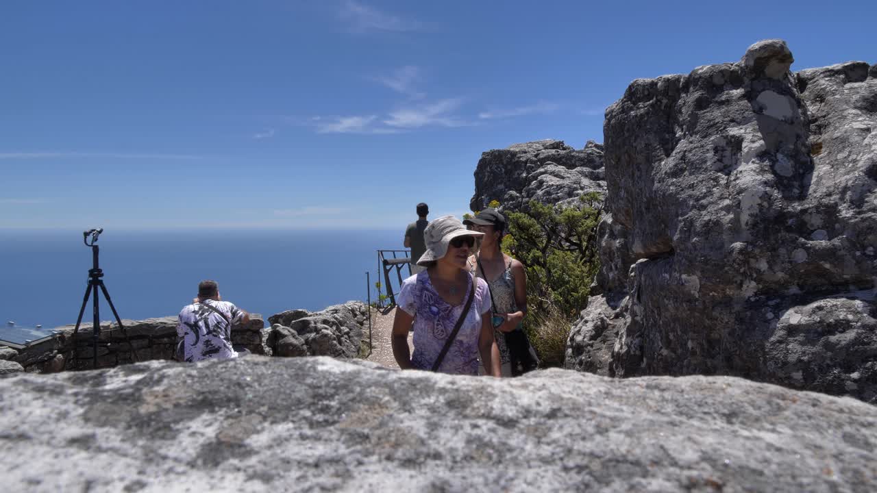 Touristen bewundern den weiten blauen Ozean und Himmel von einem erhöhten Aussichtspunkt auf dem Tafelberg, Kapstadt, umgeben von Felsformationen und Vegetation an einem sonnigen Tag.