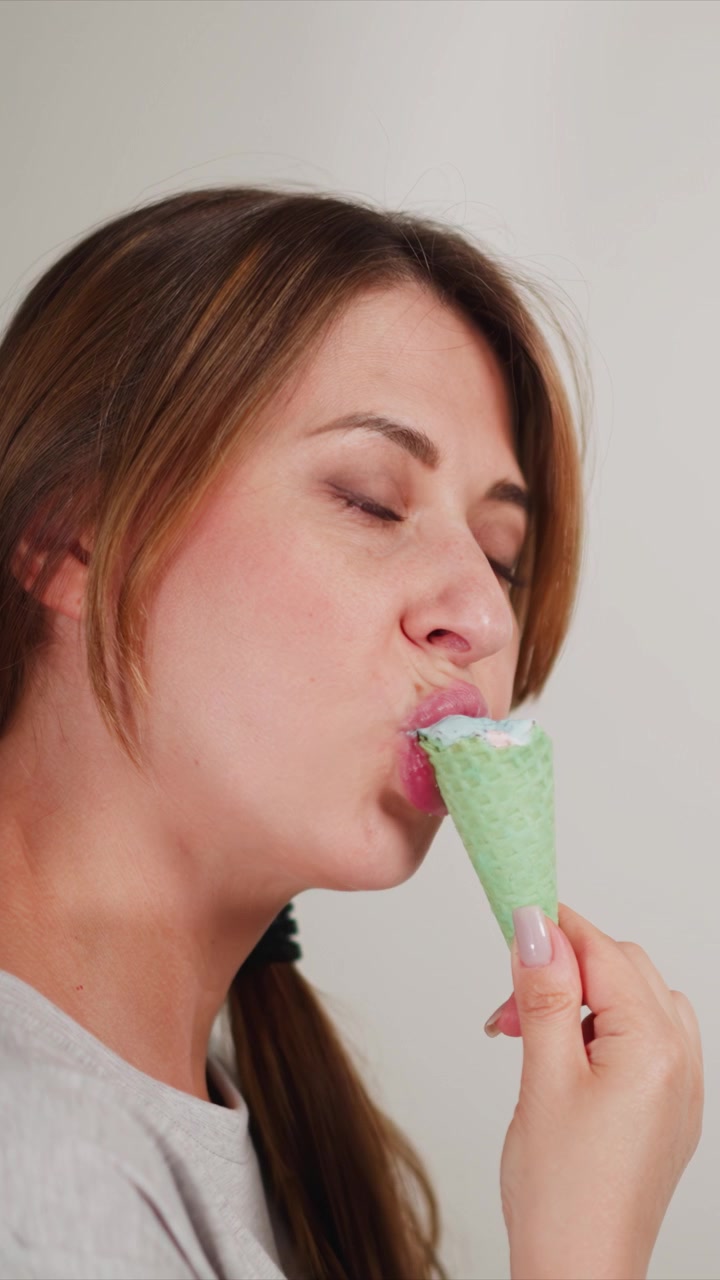 Close up of woman eating ice cream cone. subject holds green waffle textured treat with visible pink and blue swirls gradually bringing it to her mouth against plain light colored background