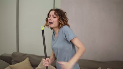 People,-housework-and-housekeeping-concept---happy-woman-in-headphones-with-mop-cleaning-floor-and-dancing-at-home.-Using-swab-pipe-as-a-microphone.-Grey-couch-on-the-background