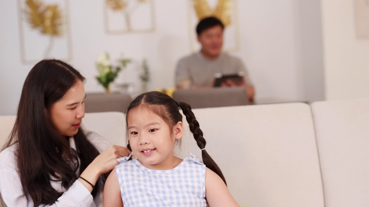 A mother braids her daughter's hair on a sofa, with a father in the background. Bright, warm lighting creates a cozy atmosphere
