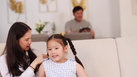 a mother braids her daughter's hair on a sofa, with a father in the background. bright, warm lighting creates a cozy atmosphere