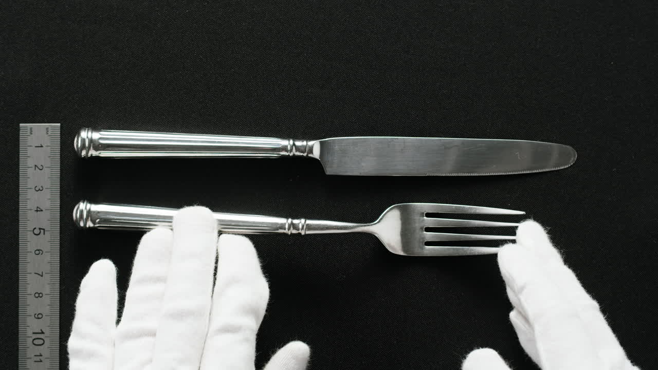 Minimalist table setting with white plate, fork, and knife arranged neatly, and a gloved hand adjusting cutlery.