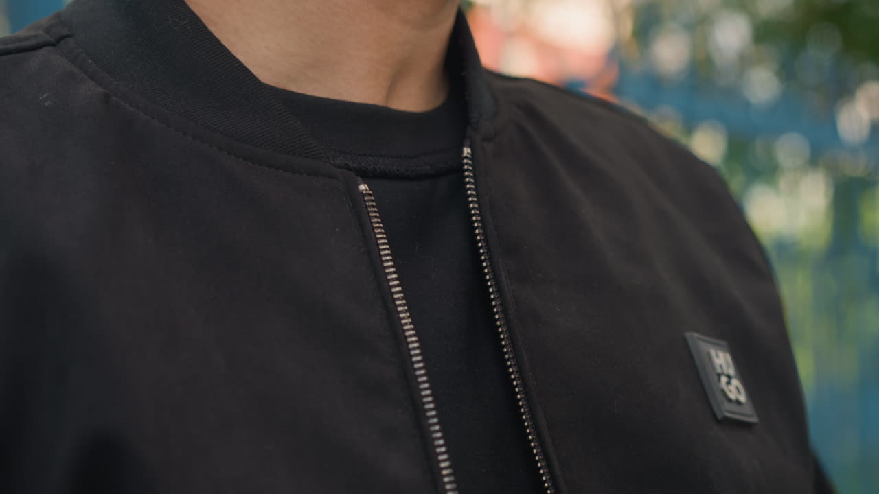 Мужчина застегивает куртку, мужчина поправляет воротник верхней одежды на улице, мужчина застегивает молнию на черной куртке на улице, крупный план мужчины, поправляющего воротник куртки-бомбера на фоне зелени