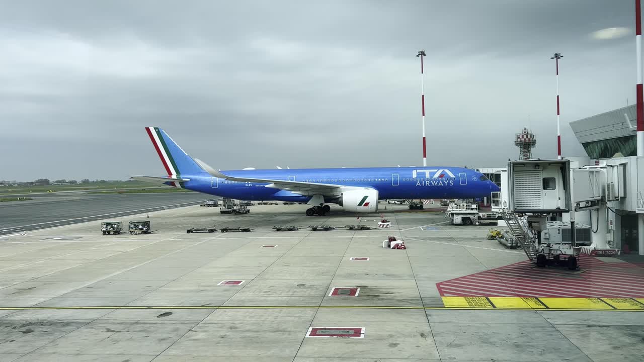 Italian Airways jet parked at boarding gate under an overcast sky, surrounded by ground service vehicles and baggage carts, showcasing air travel logistics and airport operations efficiency