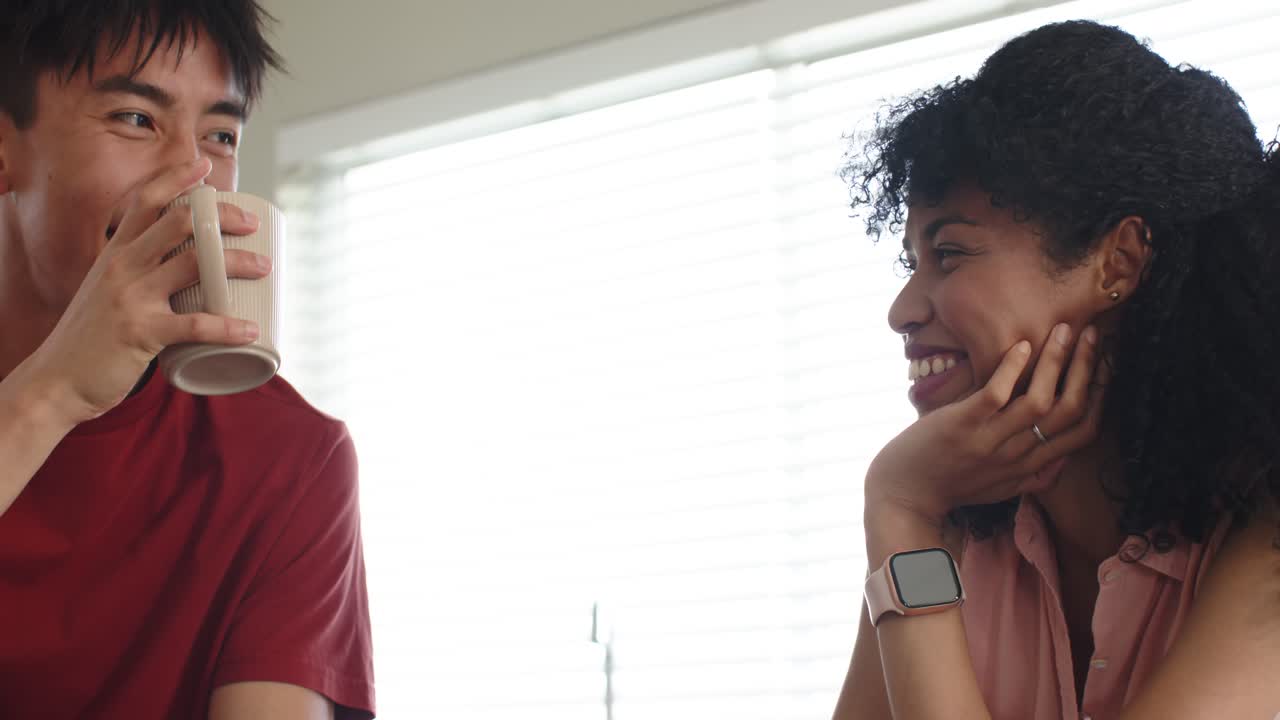 Diverse couple touching hands by mugs, laughing and sipping coffee, connecting at kitchen counter