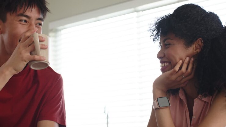 Diverse couple touching hands by mugs, laughing and sipping coffee, connecting at kitchen counter