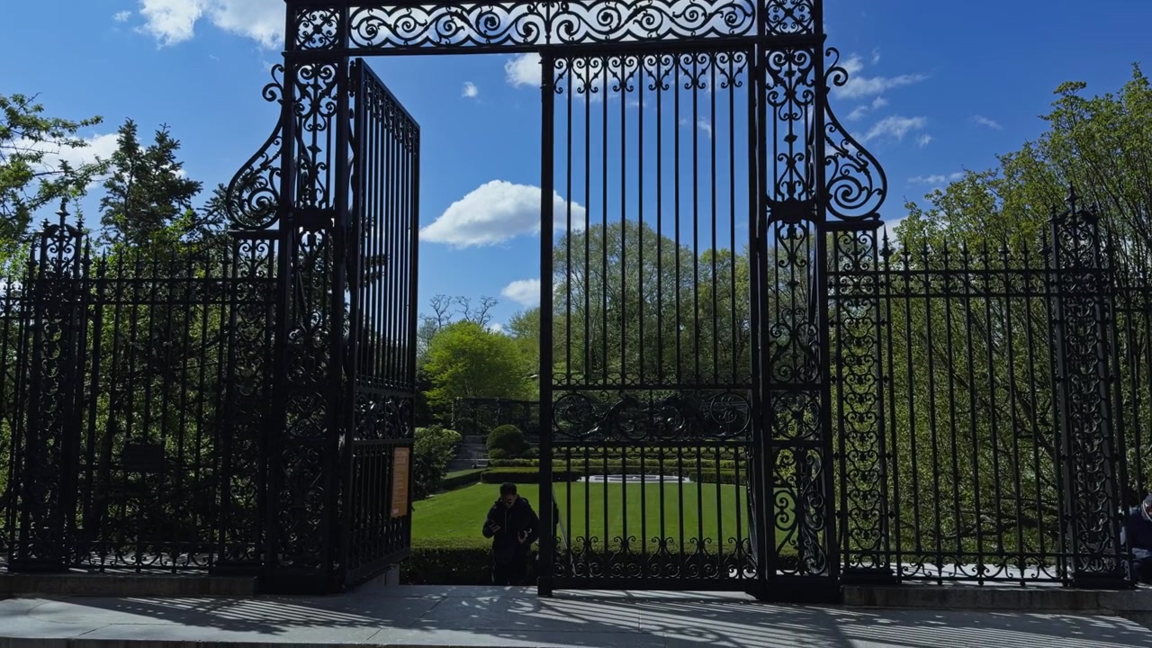 Vanderbilt Gate a Central Park, incorniciato contro un cielo azzurro e nuvole, con elaborate inferriate in ferro, inquadratura discendente che stabilisce l'ambiente.