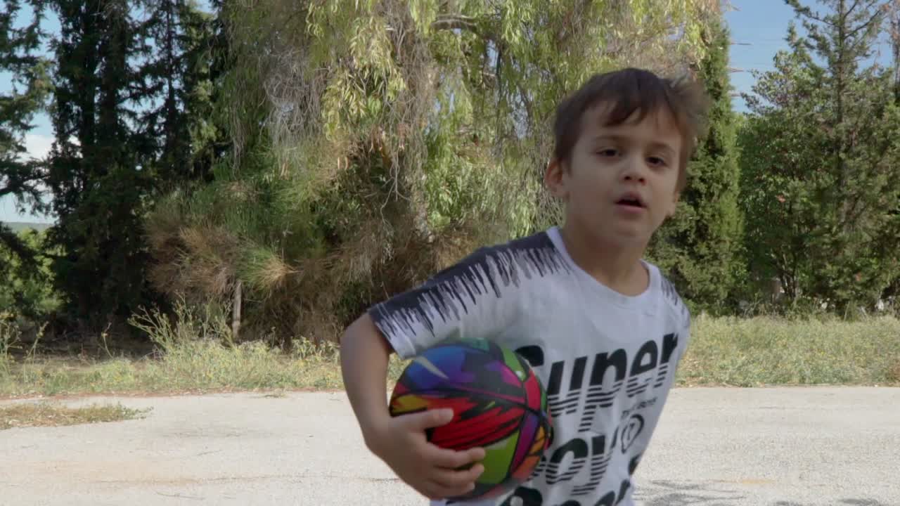Niño pequeño caucásico corriendo alrededor de un pequeño balón de baloncesto al aire libre, árboles al fondo, 120fps