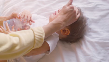 vista superior de un bebé acostado en la cama en el dormitorio mientras su madre le da agua con el biberón