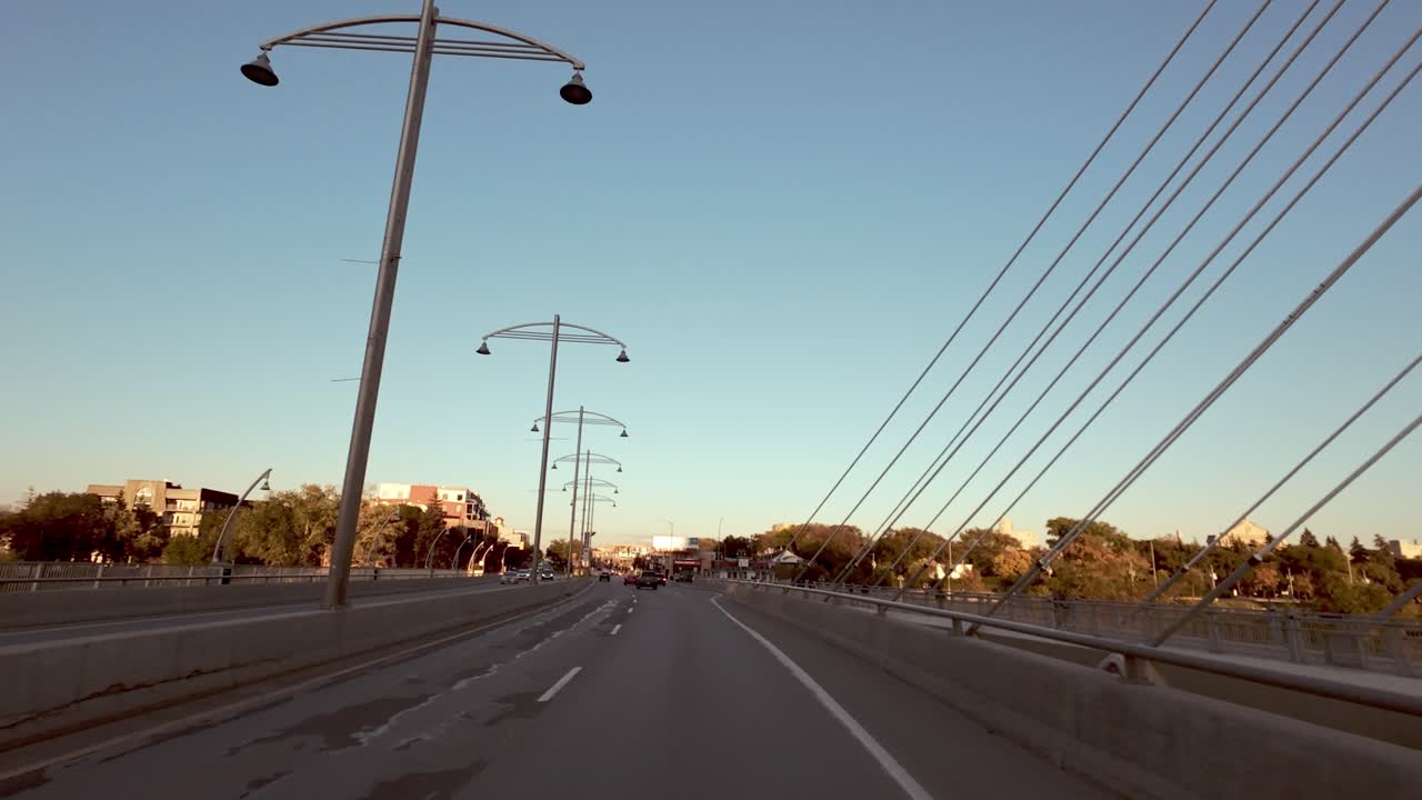 POV shot from car driving over suspension bridge with white cables; building on bridge; trees and blue sky