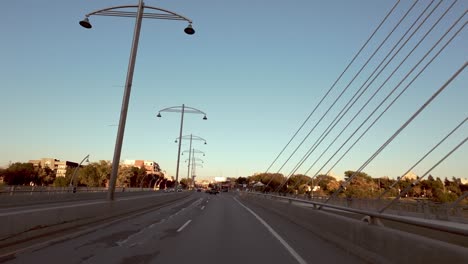 pov shot from car driving over suspension bridge with white cables; building on bridge; trees and blue sky