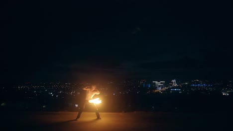 Young blond male does fire dance with two torches spins torches in the middle of the night with city skyline