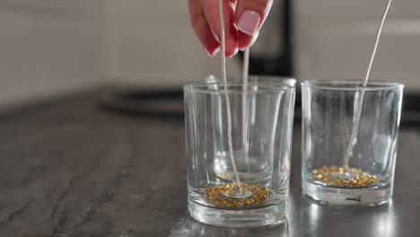 Close-up hand view of designer pouring brown beads into glass cup for creative design work, beads arranged in the cup with care, crafting process underway, organized workspace on kitchen countertop