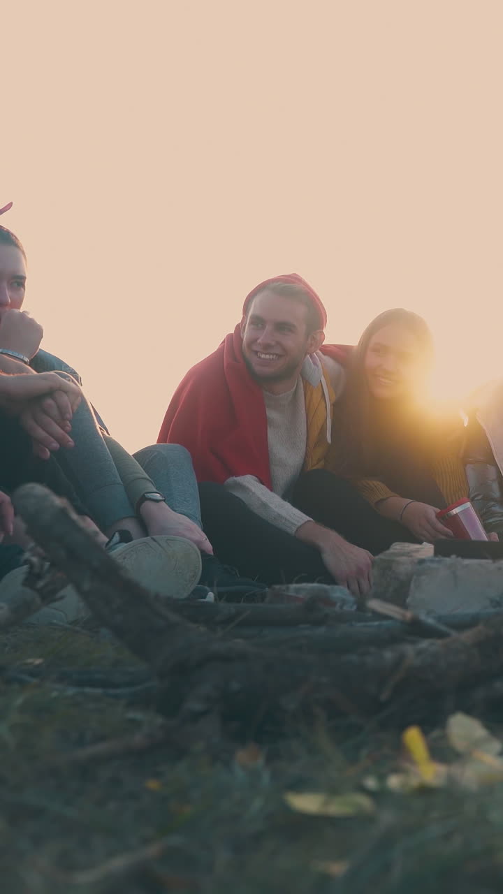 positive young friends hikers rest talking at burning bonfire on steep river bank at sunset in autumn evening