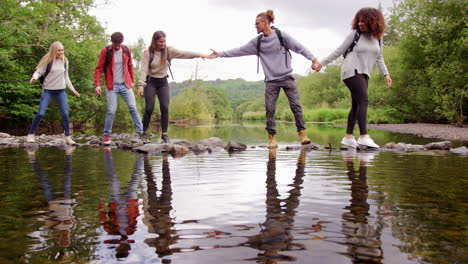 Five-young-adult-friends-hold-hands-and-help-each-other-while-crossing-a-stream-balancing-on-stones-during-a-hike,-lockdown