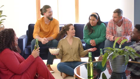 happy diverse male and female friends talking at home, drinking beers and eating popcorn