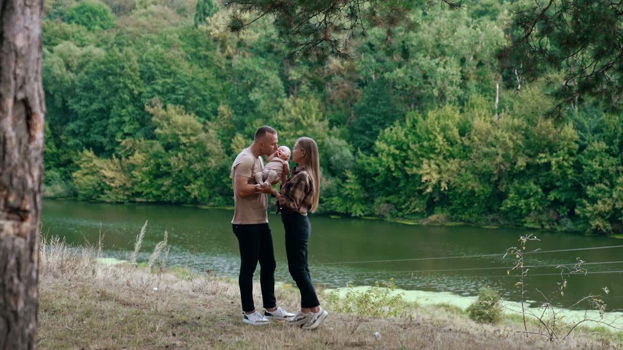 Young Caucasian parents stand at the river bank. Couple hold the infant between them and kiss the kid on the head.