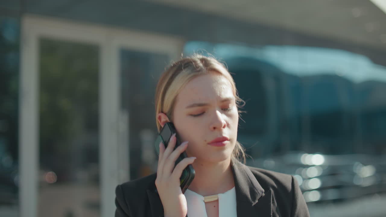 Tired woman on phone call with someone after exiting business meeting, dressed in professional attire, going home, urban glass building with sunlight on face and cars reflected in background