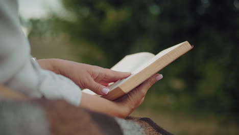 close-up of a person hands holding a book on a rustic blanket, with a softly blurred greenery background emphasizing tranquility and immersion in reading