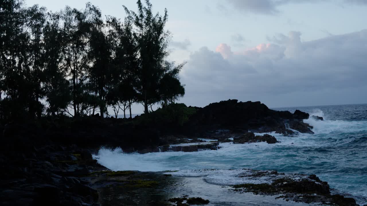 Ocean waves pound a rugged volcanic coast beneath soft twilight skies, silhouetted trees lining the shore and creating a dramatic moody atmosphere.