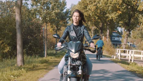 vista frontal de una hermosa y sonriente motociclista europea vestida de cuero conduciendo por carretera pasando ciclistas con árboles de color de hoja de otoño en el bosque en un día soleado