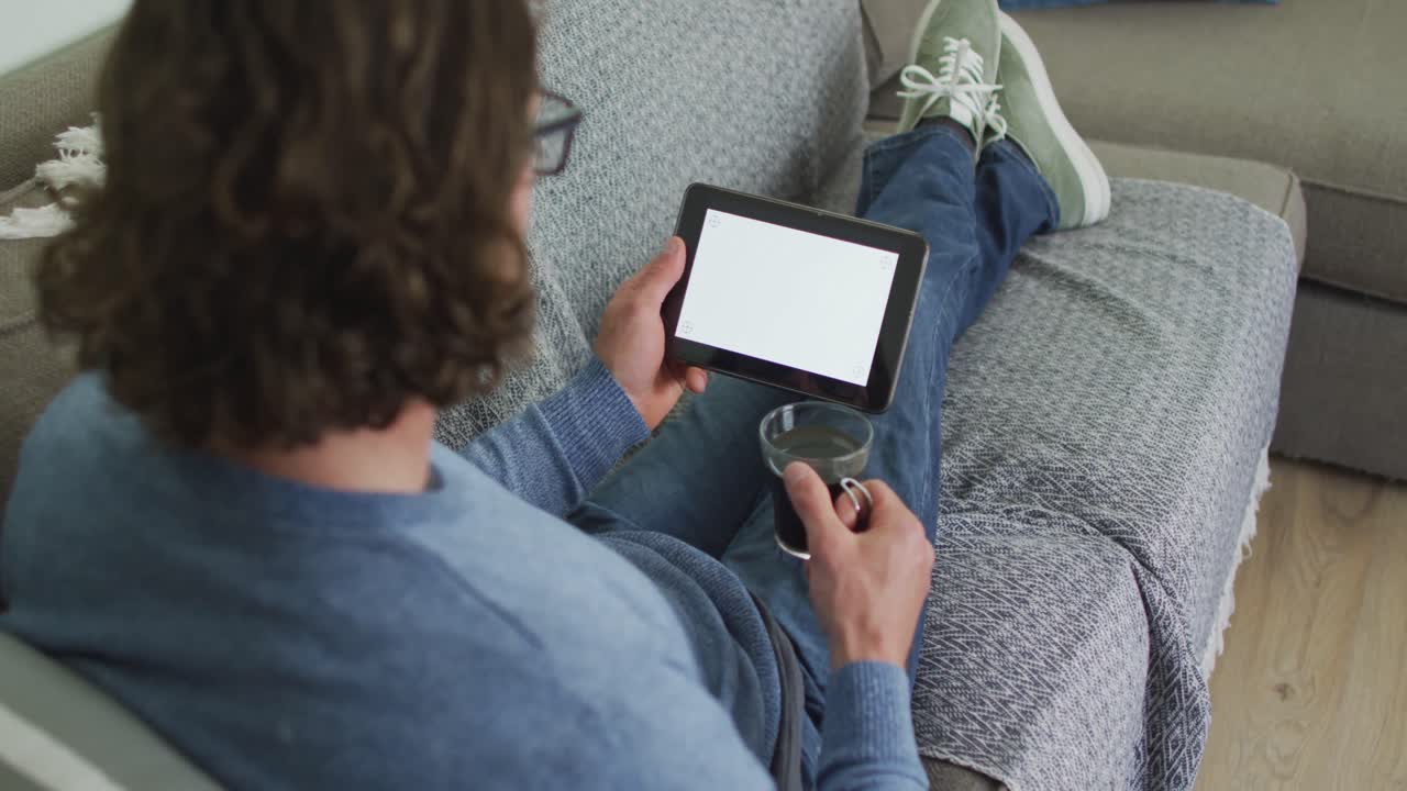 hombre caucásico sentado en el sofá en la sala de estar usando una tableta y bebiendo café