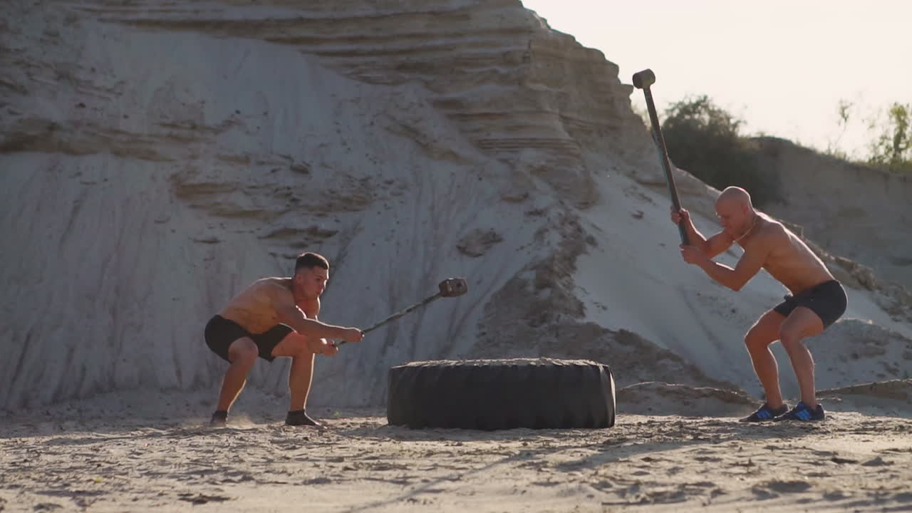 dos atletas masculinos entrenando juntos golpearon la rueda con un martillo al atardecer en las montañas en la arena. entrenamiento de resistencia