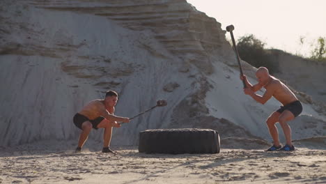 Zwei-Männliche-Athleten,-Die-Gemeinsam-Trainieren,-Schlagen-Bei-Sonnenuntergang-In-Den-Bergen-Mit-Einem-Hammer-Auf-Den-Sand.-Ausdauertraining