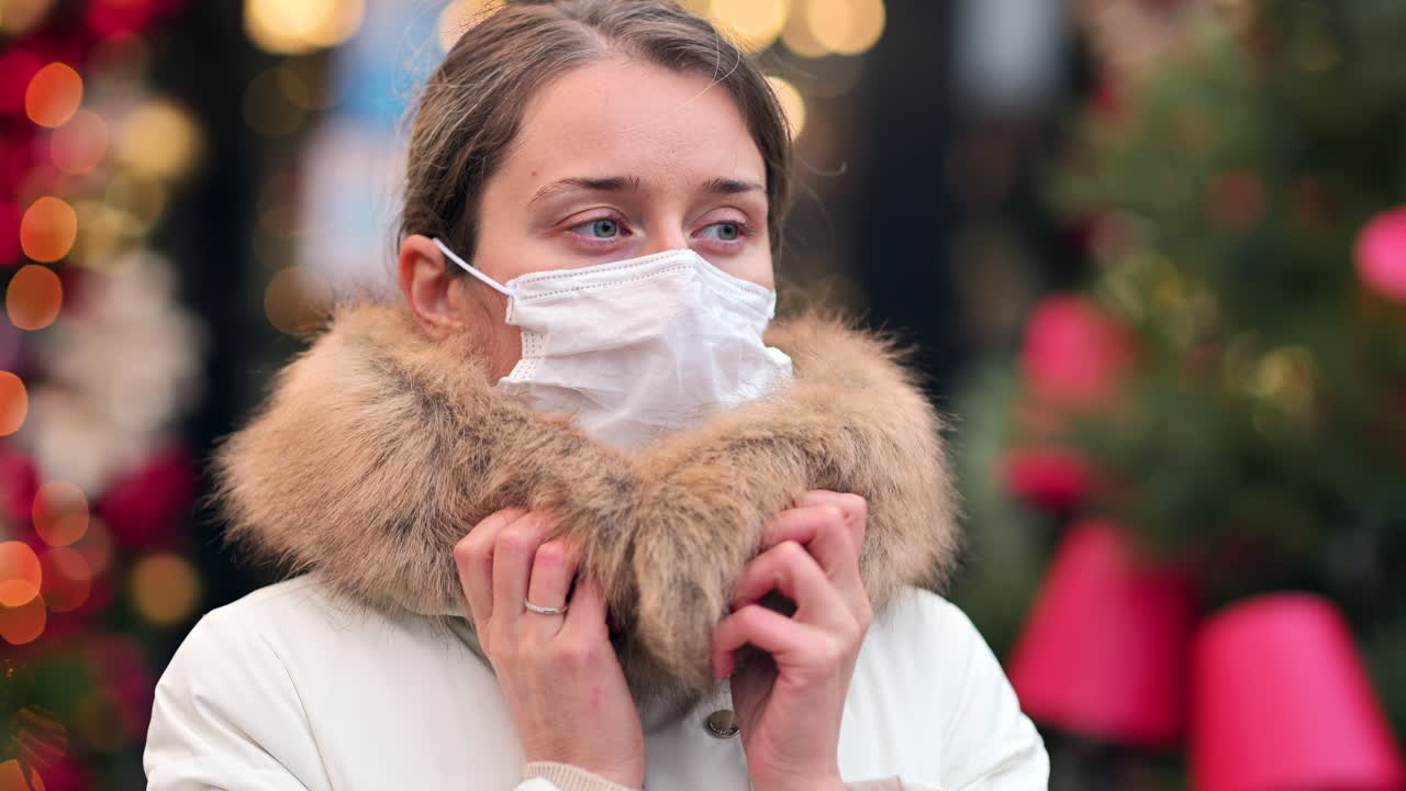 A woman puts on her face mask as she strolls through a holiday market filled with colorful lights and decorations. People around enjoy the festive atmosphere despite the chilly weather