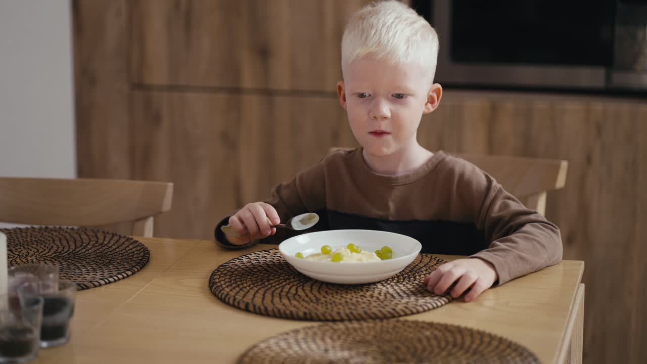 Happy little albino boy with white hair color and blue eyes eats oatmeal with green grapes while sitting on a modern chair in the kitchen. Morning before school day