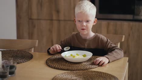 Fröhlicher-Kleiner-Albino-Junge-Mit-Weißer-Haarfarbe-Und-Blauen-Augen-Isst-Haferbrei-Mit-Grünen-Trauben,-Während-Er-Auf-Einem-Modernen-Stuhl-In-Der-Küche-Sitzt.-Morgen-Vor-Dem-Schultag