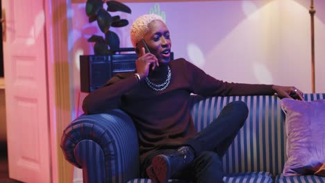 portrait of happy african american young smiling man with blonde hair talking on smartphone while sitting on a sofa in a retro room at home