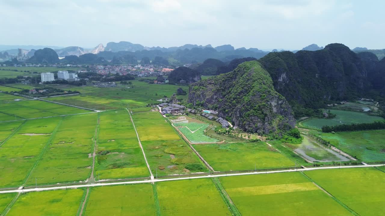 Drone pan right capturing vibrant patchwork of rice fields and steep rocky formations, with village buildings in the background