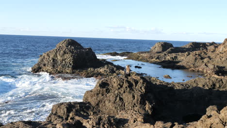 Aufnahme-Einer-Frau,-Die-Auf-Einem-Großen-Felsen-Sitzt-Und-Die-Wellen-Des-Meeres-Und-Die-Landschaft-An-Der-Küste-Der-Gemeinde-Galdar,-Auf-Der-Insel-Gran-Canaria-Und-Während-Des-Sonnenuntergangs-Bewundert