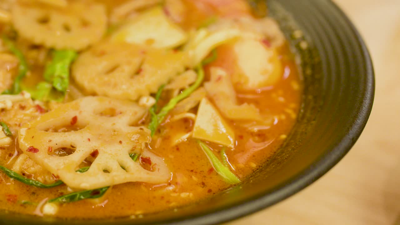 Chopsticks lift lotus root from vibrant spicy noodle soup with pork, vegetables, and hot broth