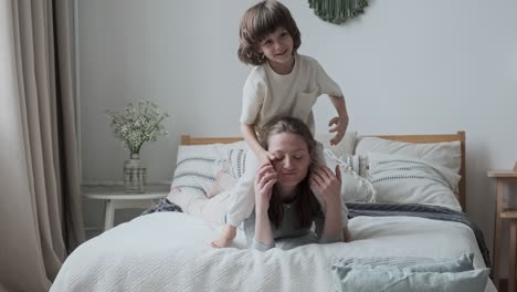 laughing male kid preschooler jumping on tired mom back sleeping on bed. family weekend activity