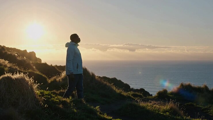 Sob o sol, um homem de jaqueta com capuz parando, levantando a cabeça, caminhando por um caminho em um penhasco costeiro, contemplando o oceano.