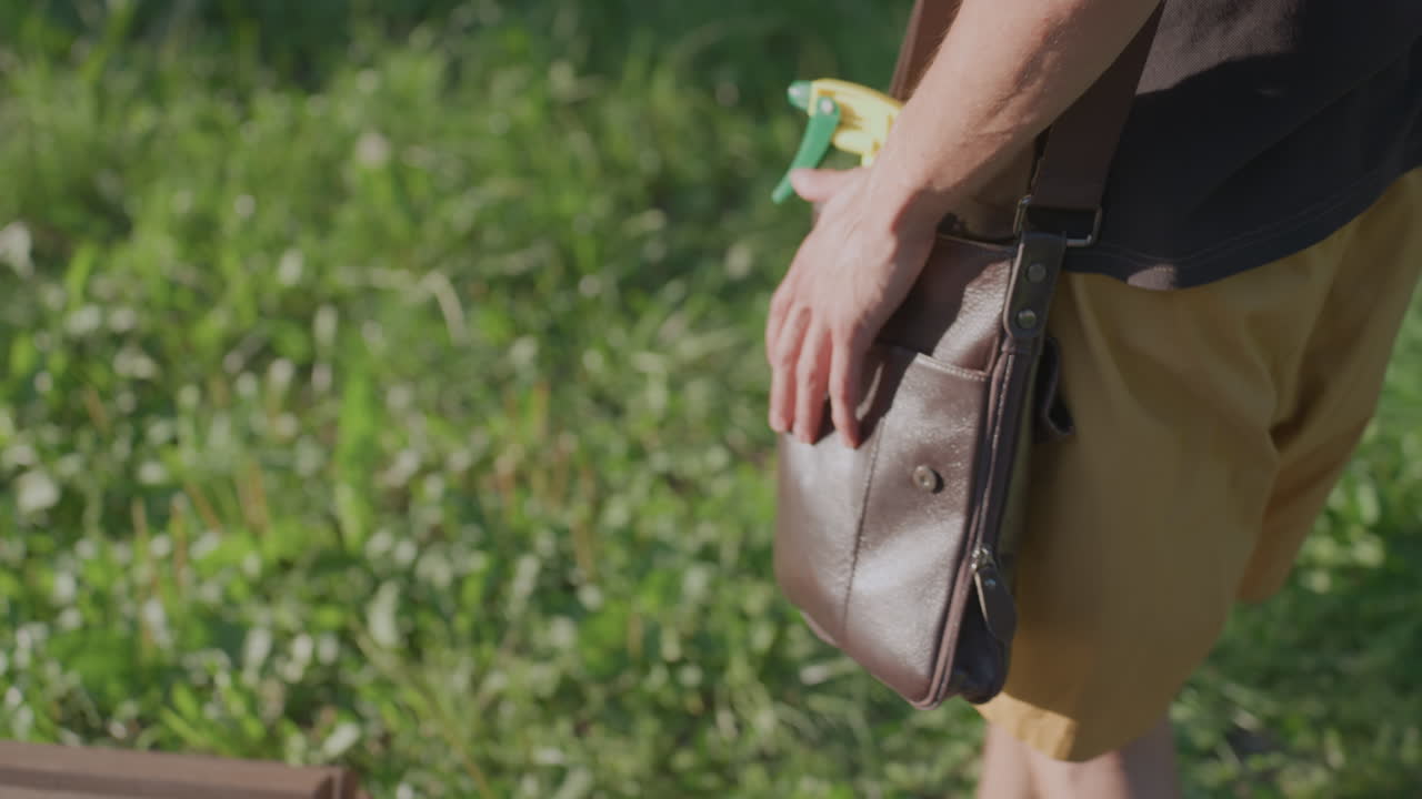 Hand Retrieves Spray, Close View Of Hand Reaching For Spray, Detailed Shot Of Hand Taking Spray From Shoulder Bag, Focused Image Of Hand Extracting Spray Bottle Amidst Grass And Bench Background