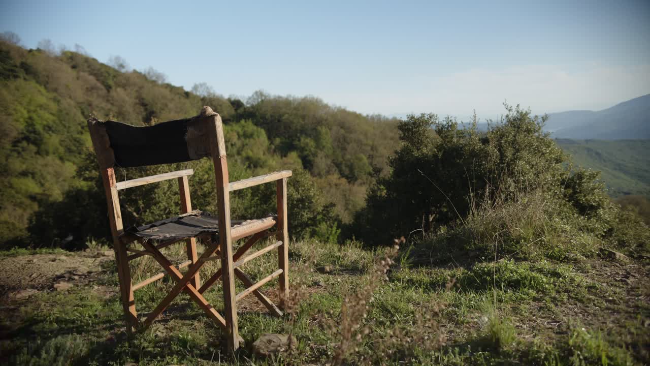 A wooden director’s chair sits empty on a grassy hillside overlooking a forested valley and distant mountains under clear blue skies. Shot in Tzoumerka.