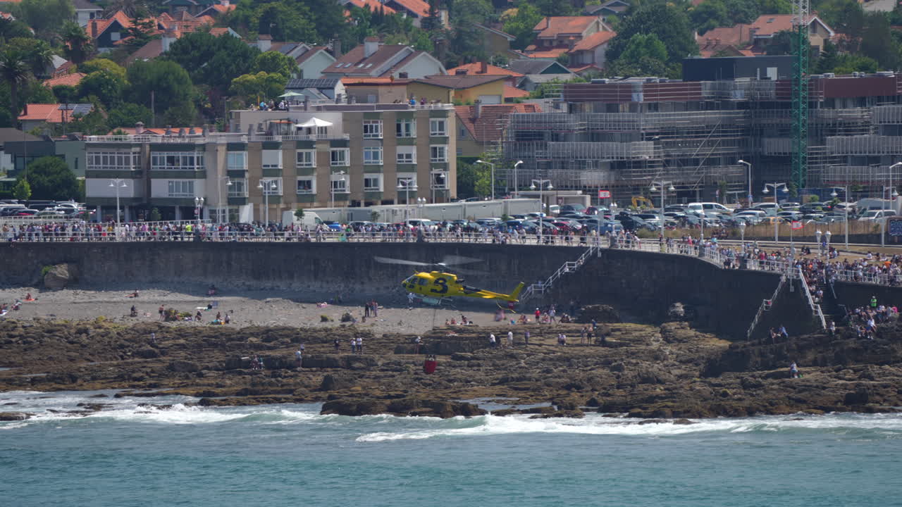 GIJON, SPAIN - JULY 27, 2025: A yellow fire helicopter lowers a water bucket near the rocky coastline of Gijon during firefighting operations. The aircraft operates close to the beach while crowds observe